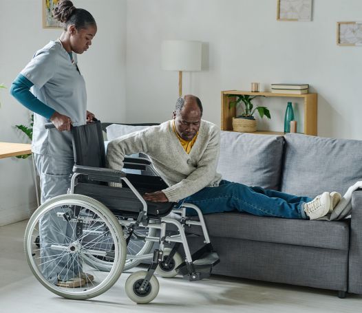 A caregiver holds the wheelchair as a man attempts to get on it from the couch.